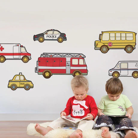 Two young children sit on a light, fluffy rug, reading books. Behind them, colorful SIMPLE SHAPES - EMERGENCY SERVICE VEHICLE REMOVABLE WALL DECALS by SIMPLE SHAPES adorn the white wall with images of a police car, school bus, fire truck, and more. The decals create an engaging and playful atmosphere that doubles as an educational learning tool.