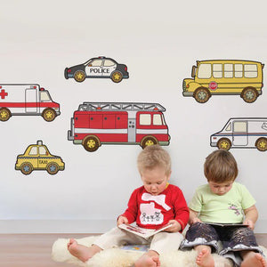 Two young children sit on a light, fluffy rug, reading books. Behind them, colorful SIMPLE SHAPES - EMERGENCY SERVICE VEHICLE REMOVABLE WALL DECALS by SIMPLE SHAPES adorn the white wall with images of a police car, school bus, fire truck, and more. The decals create an engaging and playful atmosphere that doubles as an educational learning tool.