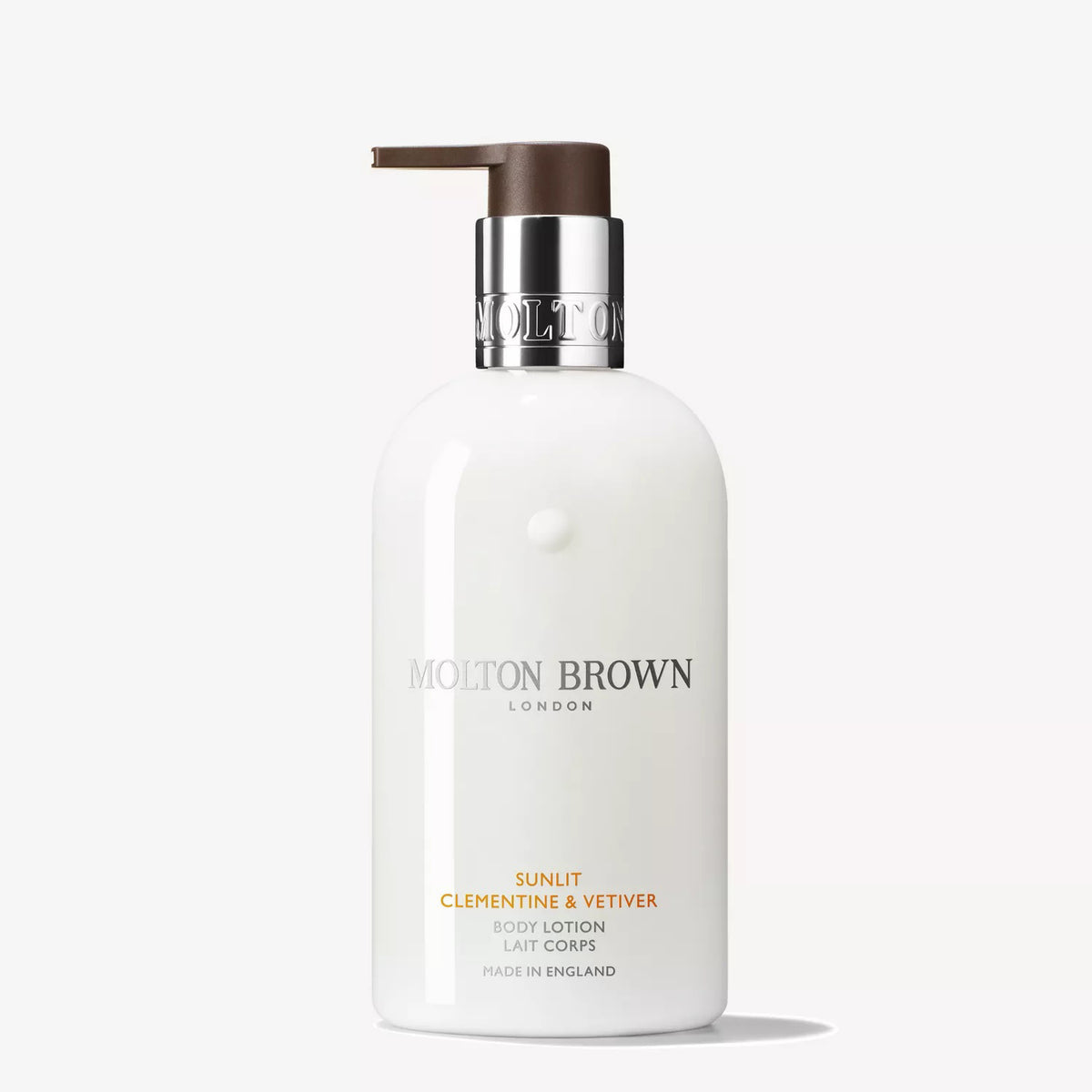 Three Molton Brown bath and body products—including the Sunlit Clementine &amp; Vetiver Body Lotion—plus a rectangular box sit on a neutral surface with a small card, set against a warm mustard-yellow background.