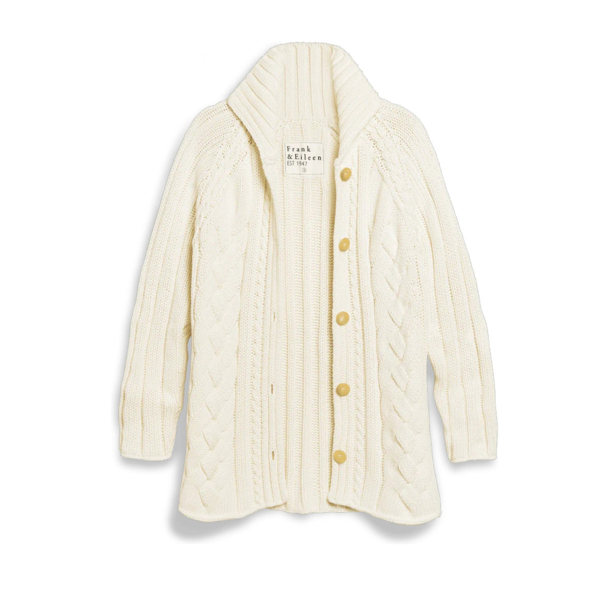 Wearing the FRANK &amp; EILEEN Cotswolds Cable Knit Cardigan in Ivory, a woman leans against a stone wall by a glass door. Behind her are shelves of vinyl records and wooden chairs; a woven bag and blanket rest on the ground nearby.
