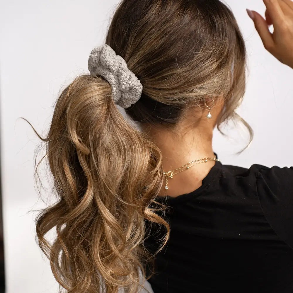 A woman with wavy, light brown hair in a ponytail secured with a VOLO BEAUTY - SCRUNCHIE DUO PACK is shown from the back, wearing a black shirt and gold jewelry, and raising her left hand.