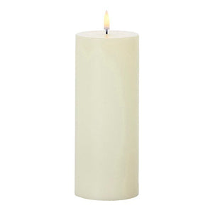 Five lit UYUNI - IVORY PILLAR CANDLES 3X8 by PIFFANY are arranged on two round wooden side tables beside a green armchair topped with a striped pillow. Tall dried grasses in a vase stand against a white wall in the background.