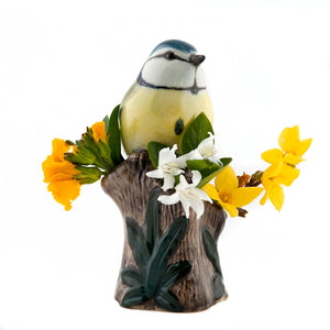 The QUAIL CERAMICS - BLUE TIT BIRD BUD VASE by QUAIL DESIGNS features a handpainted blue tit with yellow, blue, and green details perched on a brown tree stump with green leaves against a white stoneware background.