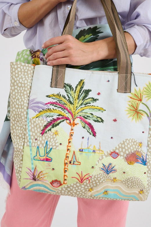 A person in pink pants, a colorful scarf, green shoes, and a light shirt holds the STORIATIPIC - CALI TOTE BAG by STORIATIPIC, an embroidered cotton bag with a palm tree and beach scene. The image is cropped above the shoulders.