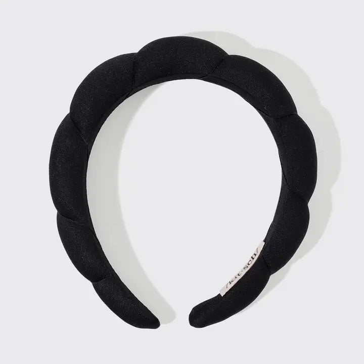 The KITSCH PUFFY HEADBAND, a black and eco-friendly accessory with a padded, segmented design, is artfully displayed against a plain white backdrop. A small tag emphasizing its sustainable nature is attached to one side.