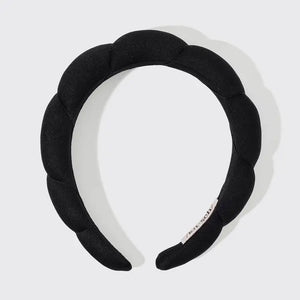 The KITSCH PUFFY HEADBAND, a black and eco-friendly accessory with a padded, segmented design, is artfully displayed against a plain white backdrop. A small tag emphasizing its sustainable nature is attached to one side.