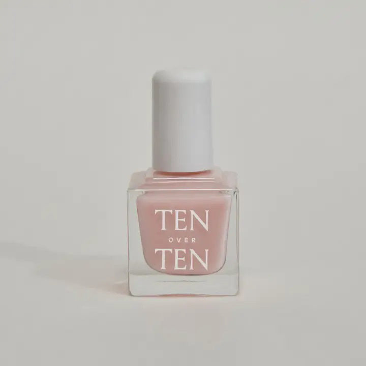 A glass bottle of TENOVERTEN - NAIL POLISH from the brand TEN OVER TEN, featuring a white cap and a dusty pink nail color. The front label displays "TEN OVER TEN" in white uppercase letters. This vegan, eight-free formula ensures both beauty and peace of mind. The background is plain and light-colored.