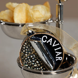 A ZODAX GLASS CAVIAR BOX ORNAMENT shaped like a 3-inch round tin with “CAVIAR” text and metallic beads, sitting in a silver serving dish next to a bowl of potato chips.