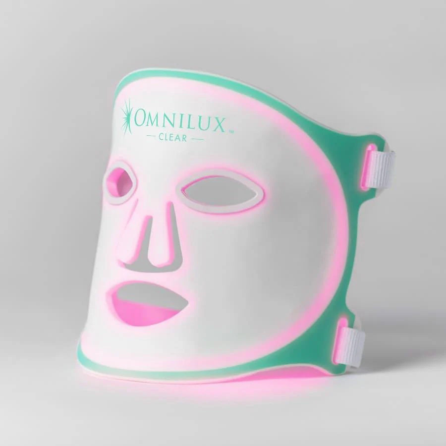 The OMNILUX CLEAR MASK, offering white and teal LED light therapy for acne with eye, nose, and mouth cutouts, uses blue and red clinically proven light therapy. It includes a controller, charging accessories, and an instruction manual.