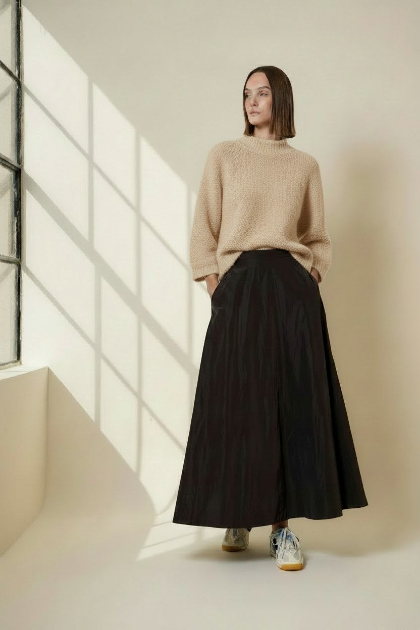 A woman stands indoors by a window, wearing the VICARIO CINQUE - MADEIRA Boucle Sweater in Light Taupe from VICARIO CINQUE, paired with a long black skirt and sneakers. Sunlight casts grid-like shadows on the wall.