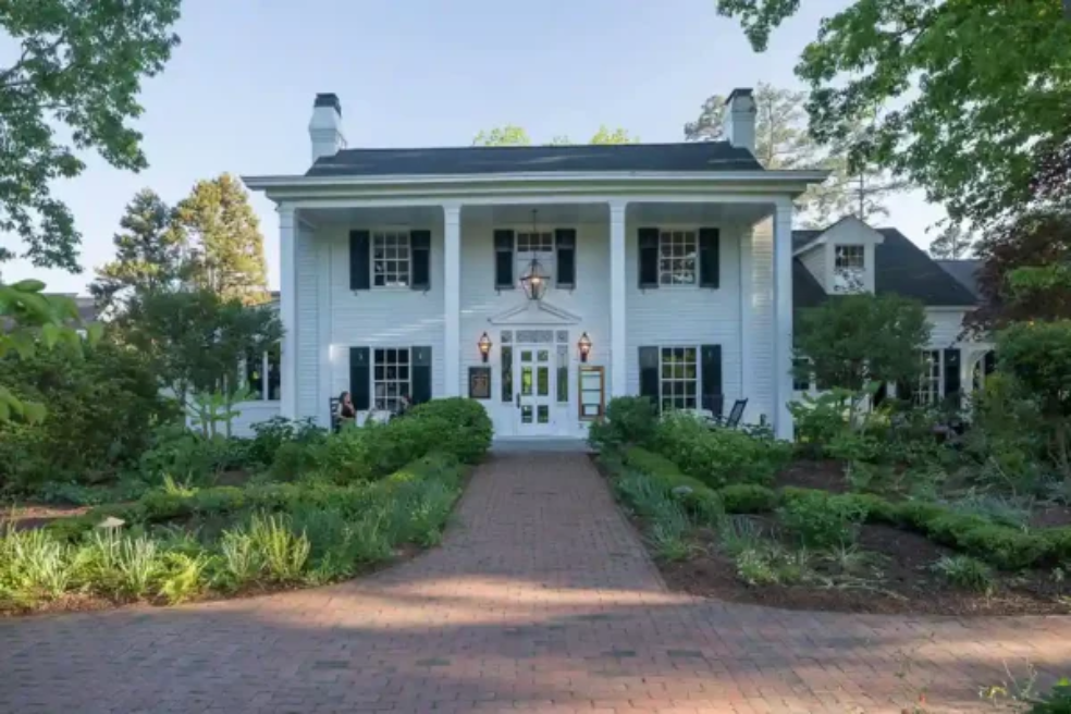 A large white two-story house with black shutters and columns, a brick walkway leading to the front door, and lush green landscaping surrounding the entrance.