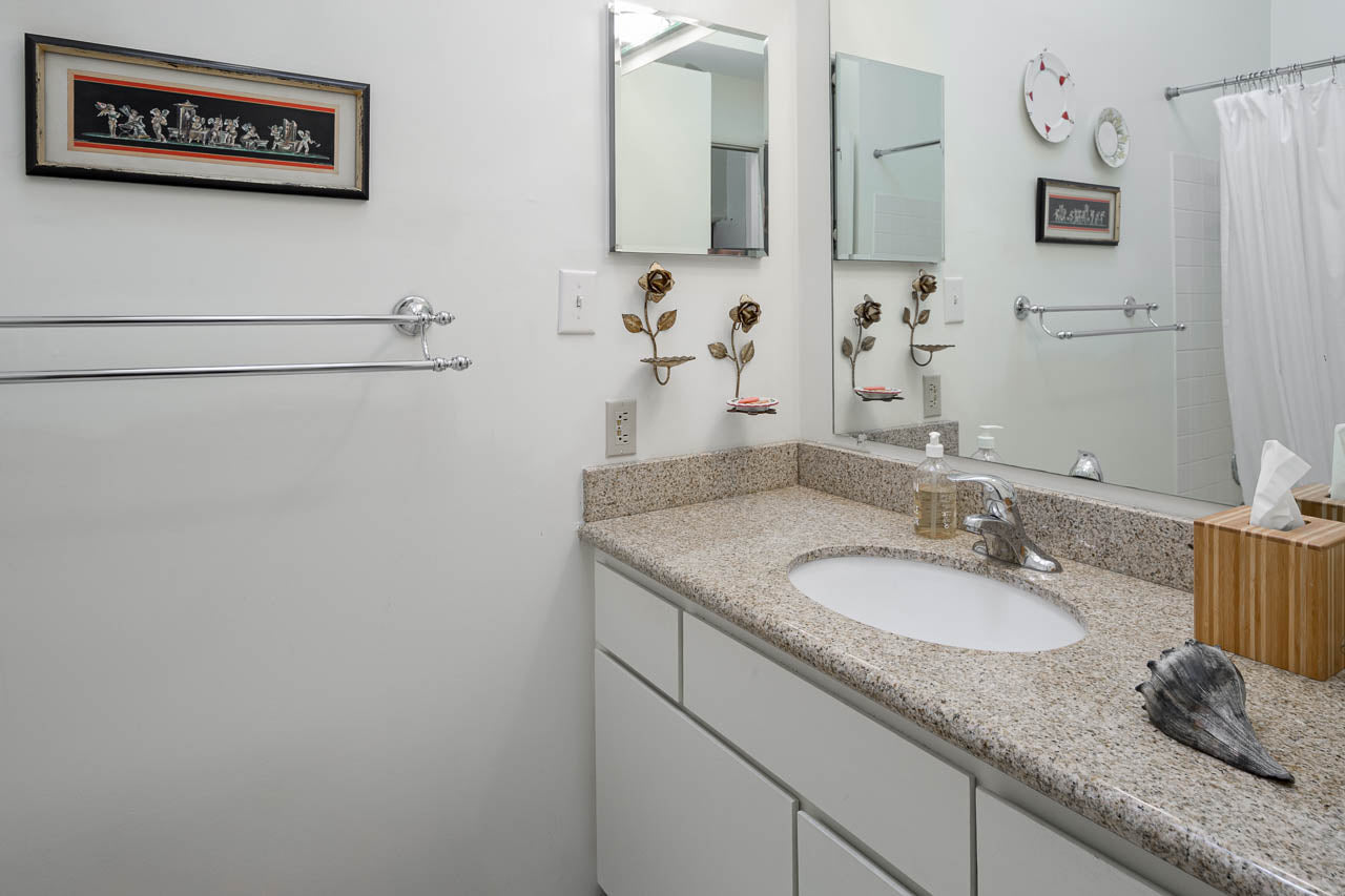 A clean bathroom with a granite countertop, white sink, mirrors, towel racks, a tissue box, soap dispenser, seashell, and wall art above the counter. The shower with a white curtain is visible in the background.