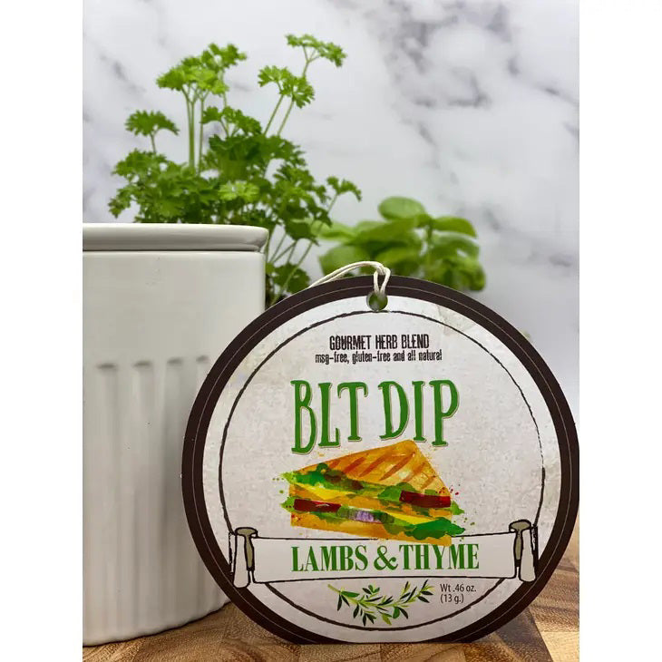 Close-up of a round package labeled &quot;LAMBS &amp; THYME - BLT DIP&quot; with an illustration of a sandwich featuring bacon and tomato. The label indicates it is a gourmet herb blend by Lambs &amp; Thyme. A white container and green leafy plant are in the background on a light-colored countertop.