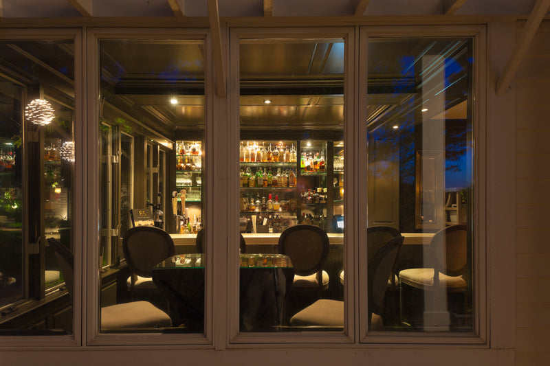 A view through large windows into a warmly lit bar with a fully stocked liquor shelf, elegant chairs, and a round table, set against a dark evening background.