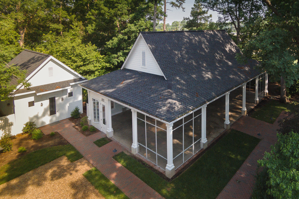 Aerial view of a white house with a screened-in porch and a nearby detached garage, both surrounded by trees, brick walkways, and green lawn. Sunlight filters through the branches, creating dappled shadows.