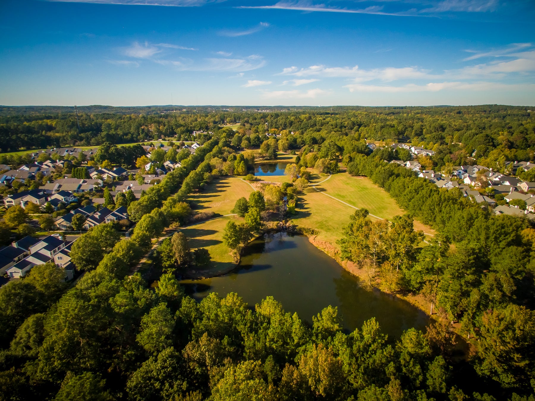 Aerial view of a suburban neighborhood with houses, green lawns, trees, and several small ponds under a blue sky with scattered clouds.