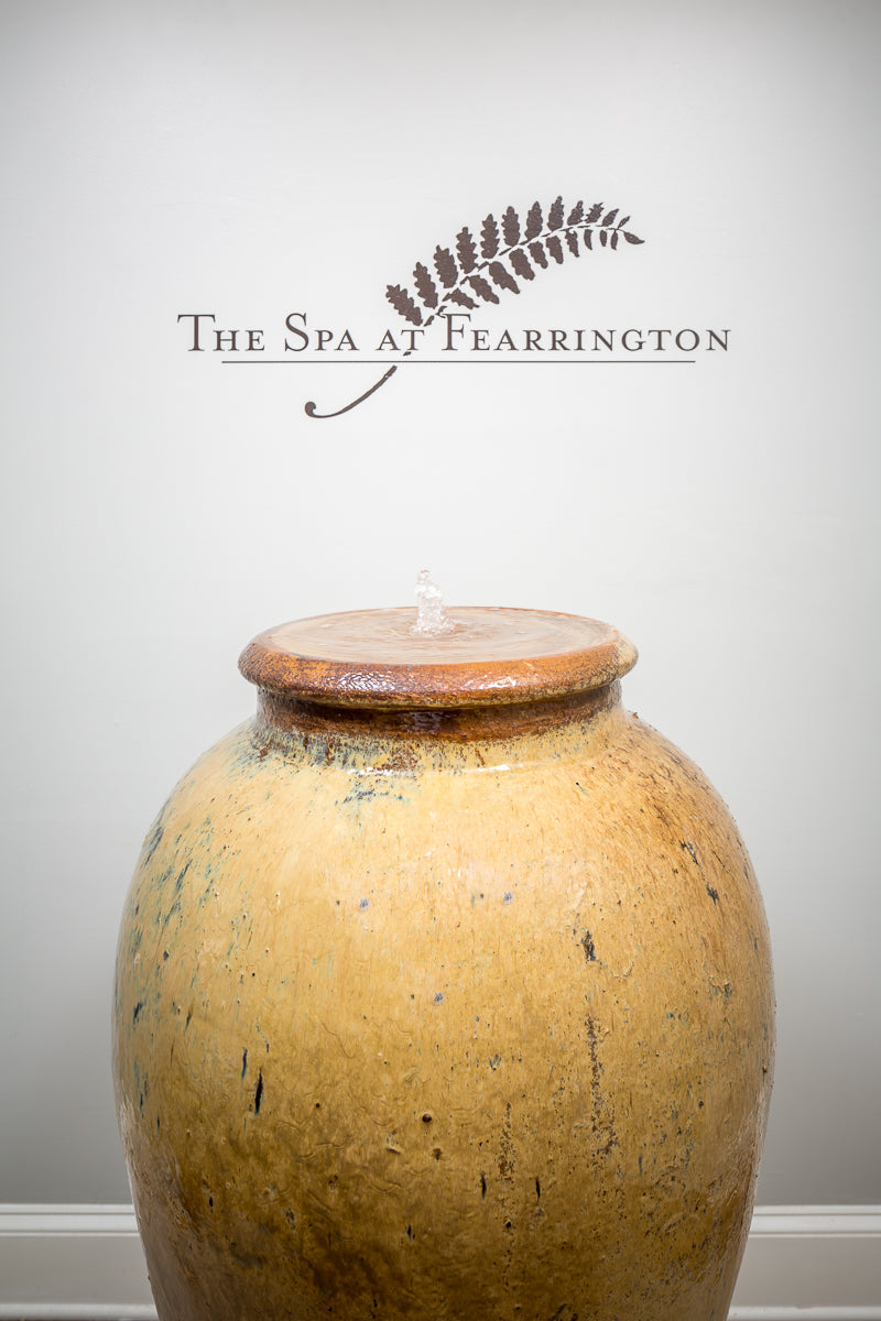 A large ceramic water jar with a small fountain on top sits in front of a white wall with 