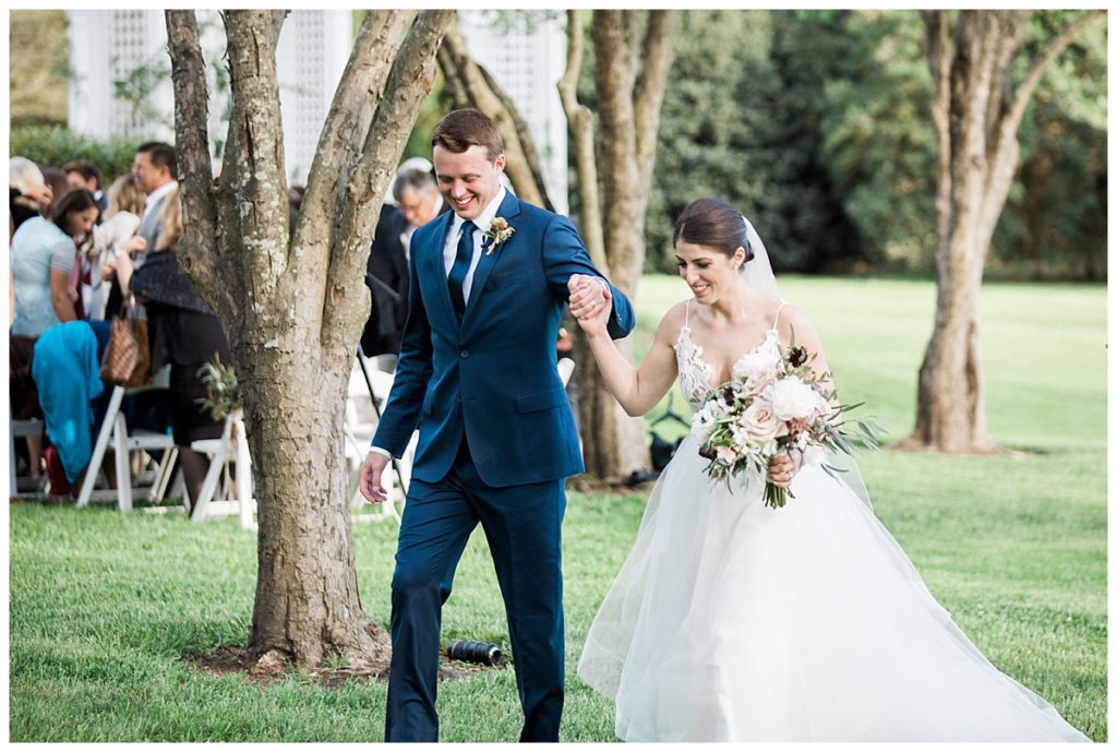 A smiling bride and groom walk hand in hand outdoors, surrounded by trees and greenery. The bride holds a bouquet of flowers and wears a white dress, while the groom wears a blue suit. Guests are seated in the background.