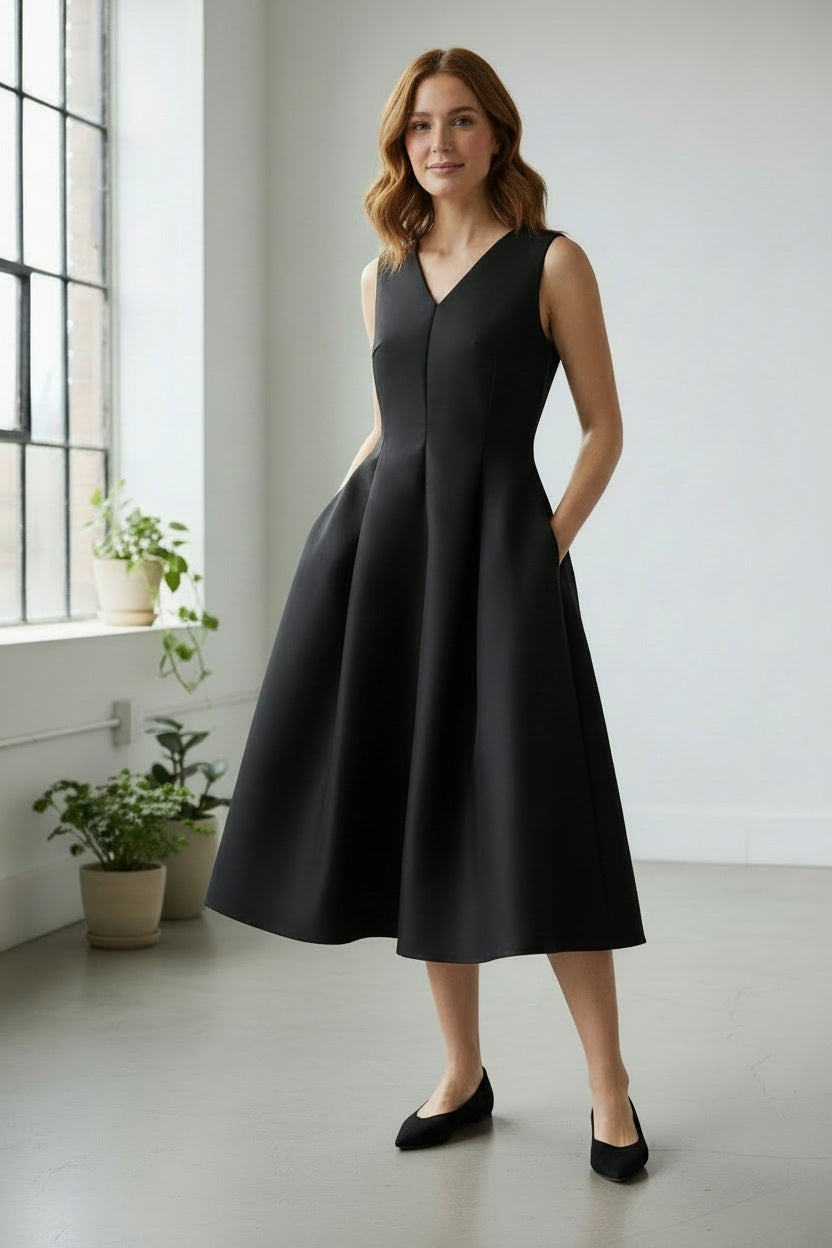 A woman wearing the PARVATI DRESS IN BLACK by ALYKI LABORATORIO stands in a bright room with large windows and potted plants, her black flats complementing the elegant A-line midi dress with pockets as she smiles softly.