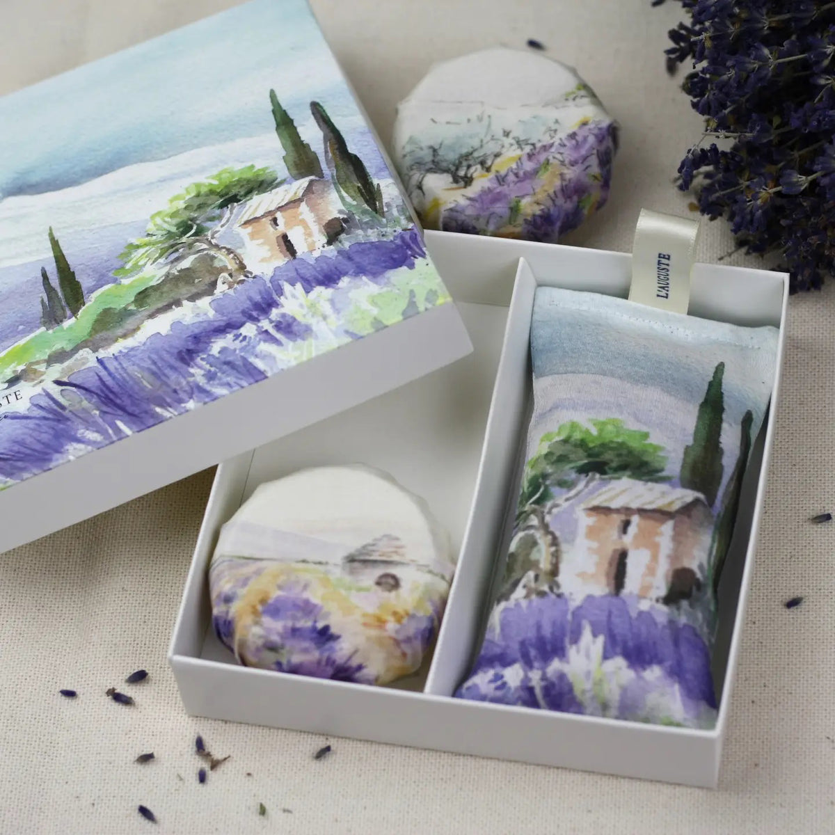 The L&#39;AUGUSTE PROVENCE - SUMMER IN PROVENCE Lavender Sachet and Soap Gift Box by L&#39;AUGUSTE PROVENCE features organic lavender soap and a sachet with watercolor art of a Provencal house, trees, and fields. Purple lavender flowers sit beside the box.