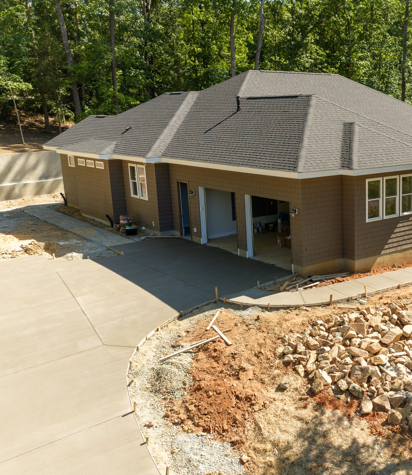 A new concrete driveway curves toward a modern brown house with a two-car garage, surrounded by trees and landscaping with rocks and soil.