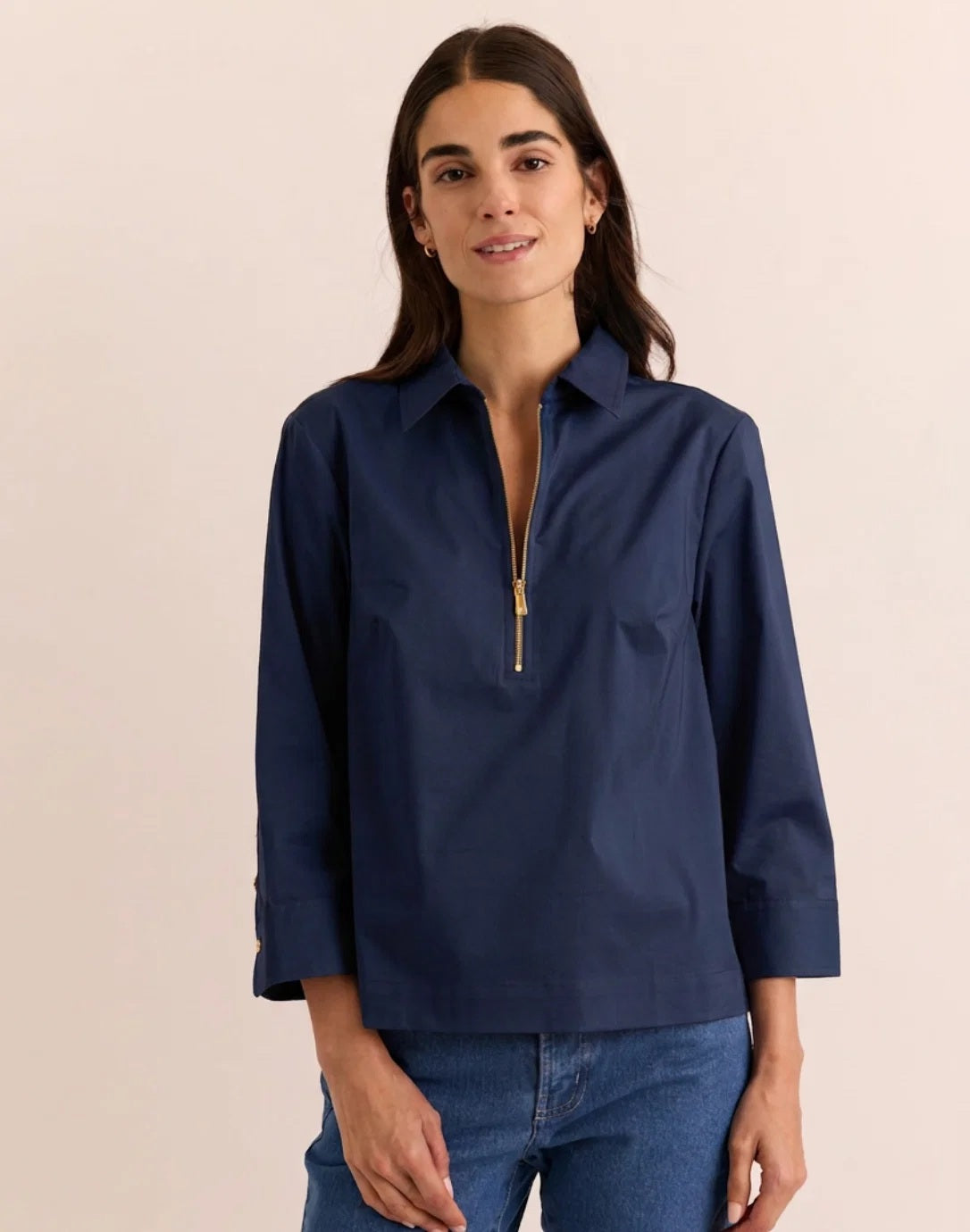 A woman with long dark hair models the HINSON WU Alessa 3/4 Sleeve Shirt in Stretch Polished Cotton Navy, featuring a collared neckline and gold zipper detail, paired with blue jeans against a plain beige background.