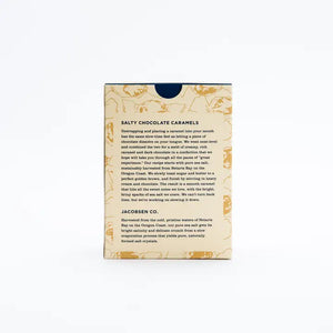 A rectangular box of JACOBSEN SALT CO. CHOCOLATE CARAMELS WITH SEA SALT stands on a light surface, featuring tan packaging with brown abstract patterns and a white label displaying the product name in black and gold.