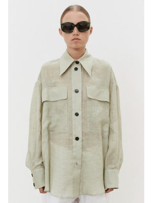 A person with brown hair in a low ponytail faces away, wearing an oversized MEIMEIJ - LINEN SHIRT. Against a plain white background, breathable linen pants are visible at the bottom.