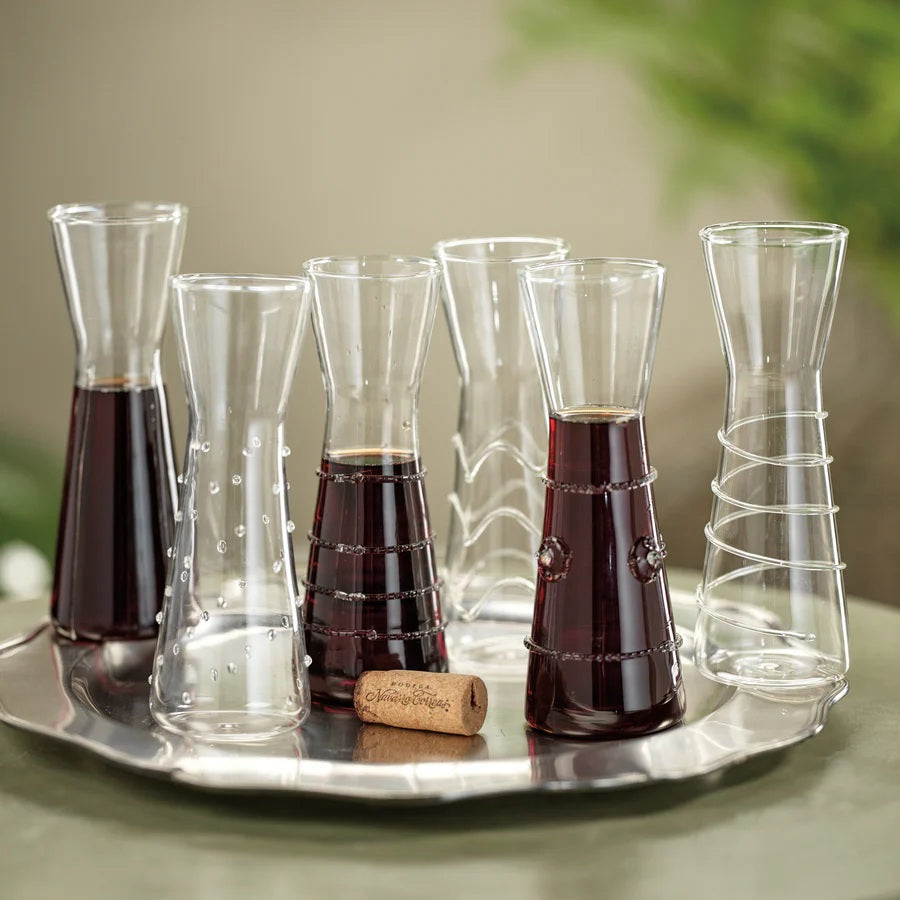Six ZALLI SINGLE SERVE GLASS CARAFES by ZODAX, each featuring a unique etched design, are arranged on a reflective silver tray with a blurred green background.