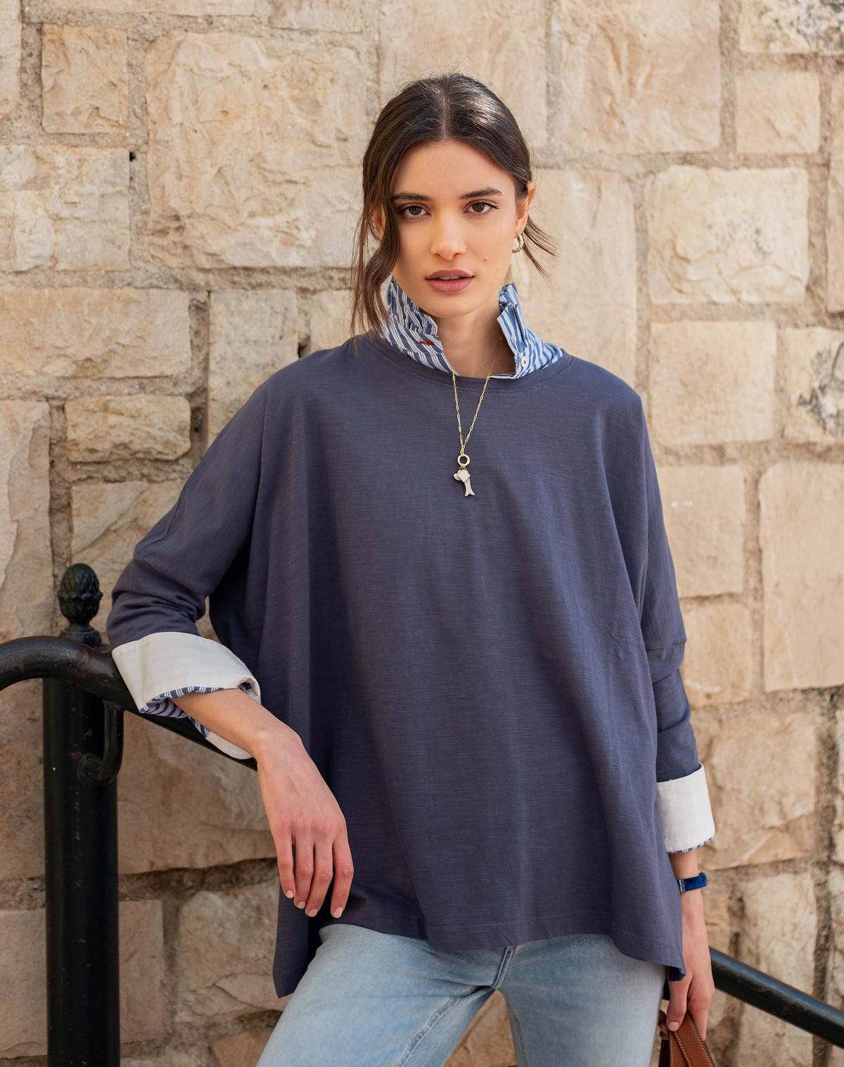 A young woman stands in front of a stone wall, looking at the camera, with her light brown hair tied back. She is wearing a necklace, light blue jeans, and the MERSEA - Catalina Slub Tee in Indigo—a relaxed top from MERSEA featuring 3/4 length sleeves with rolled cuffs. The soft cotton-blend slub fabric gives her look an effortlessly stylish and comfortable vibe.