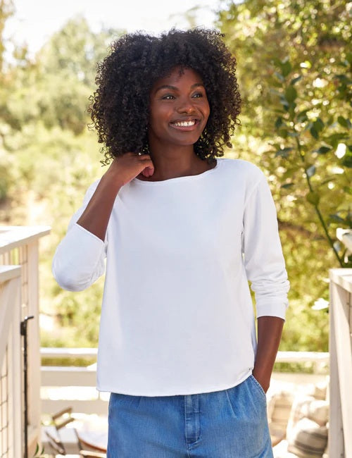 A woman with curly hair smiles outdoors, wearing the FRANK & EILEEN Ella Long Sleeve Boatneck Tee in Heritage Jersey White from the California Heritage Collection, and displaying a ring on her left hand.