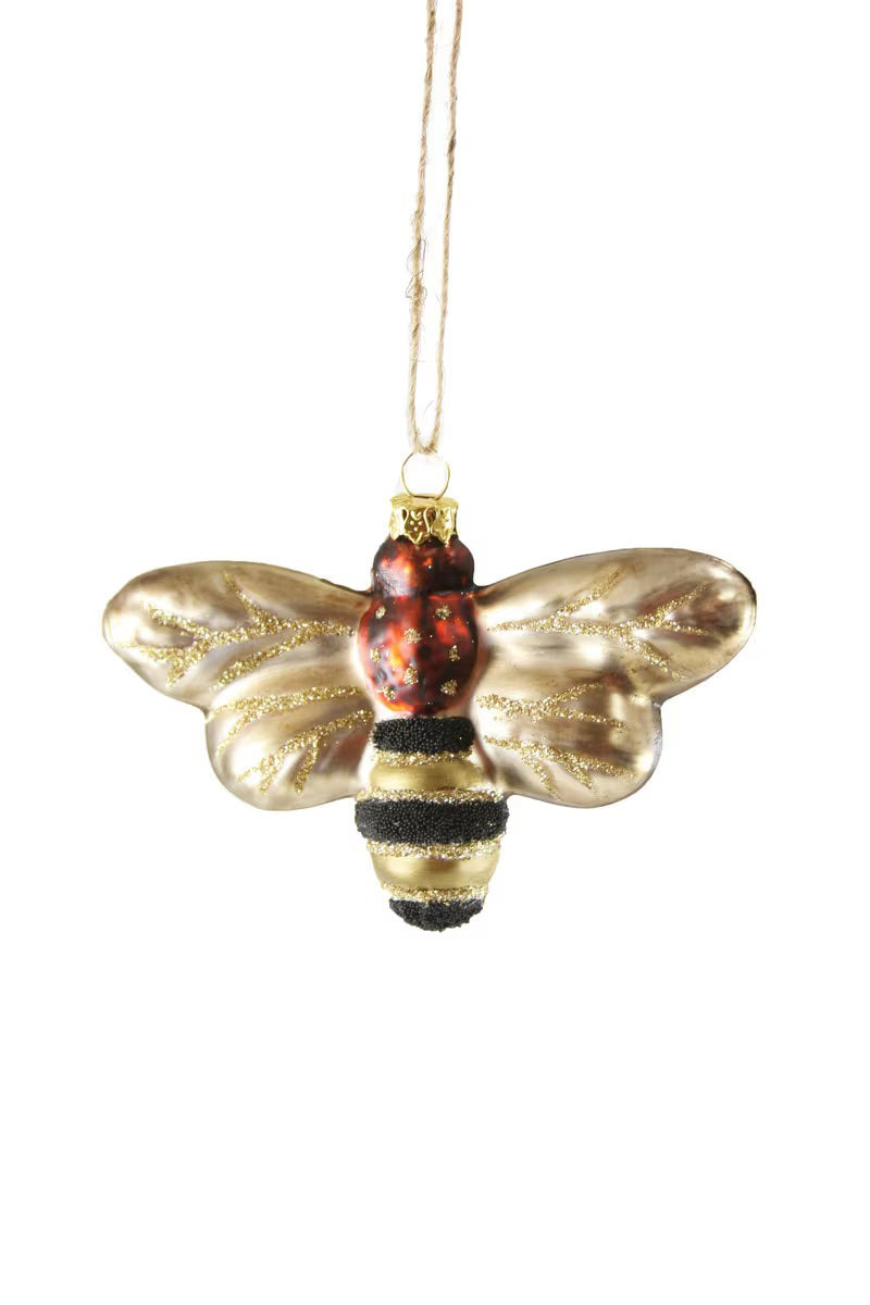The HONEY BEE ORNAMENT by CODY FOSTER is a decorative piece featuring a bee shape with glittery wings and a glass finish, showcasing a red and black body along with a gold-striped abdomen. It hangs intricately on a string against a white background.
