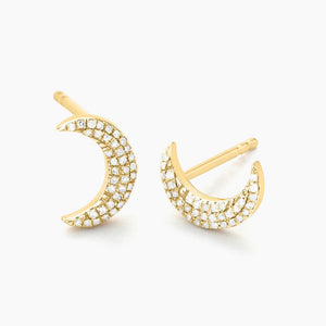 A close-up of a woman’s ear wearing the ELLA STEIN - MINI MOON STUD EARRINGS by ELLA STEIN, featuring small crescent moon shapes adorned with sparkling natural diamonds set in gold-plated stainless steel. Brown hair with blonde highlights is visible in the background.