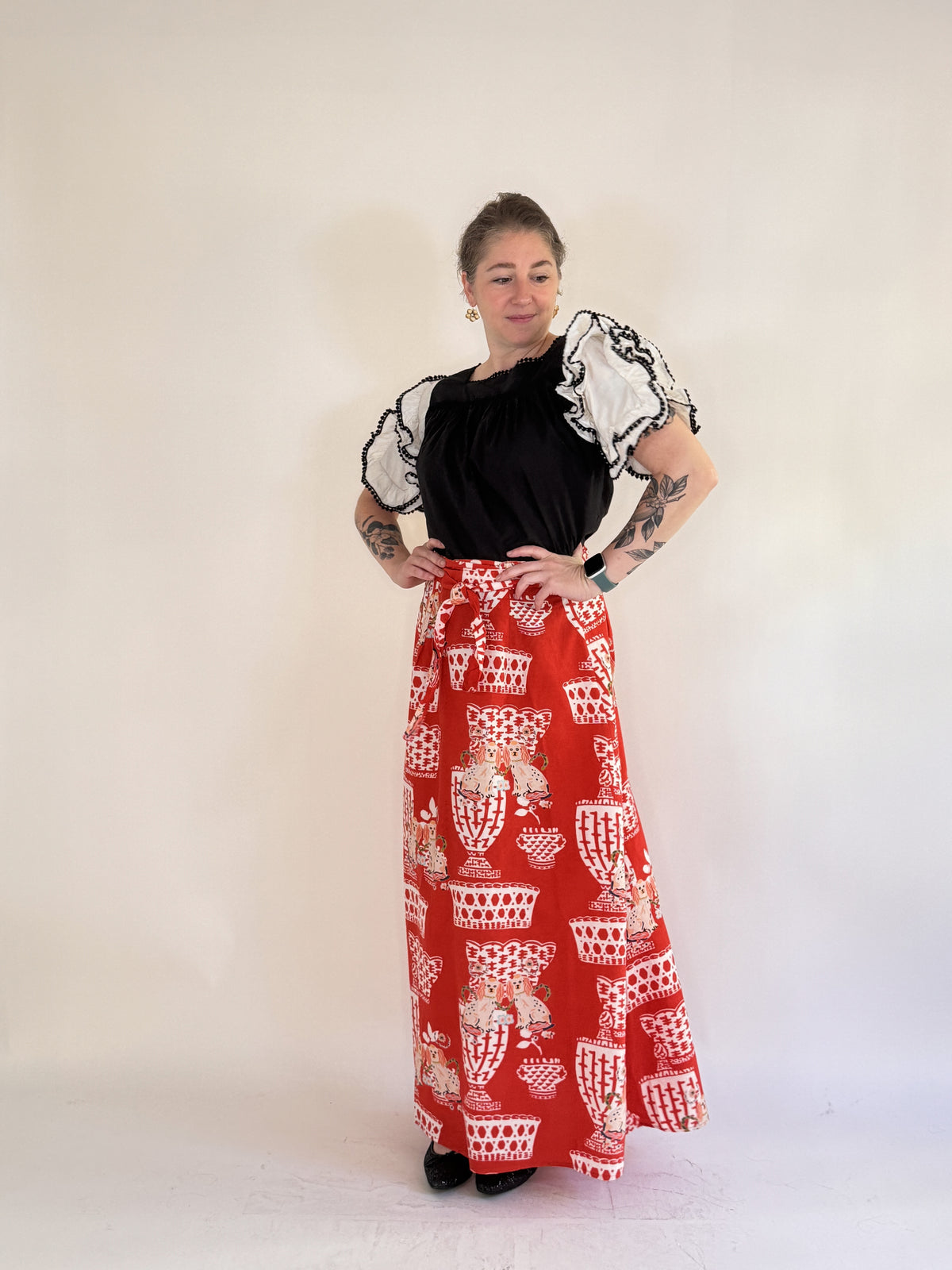 A person with tattooed arms stands against a light background, wearing a black top with sheer ruffled sleeves, black shoes, and the FARYLINA - FIFI WRAP SKIRT by FARYLINA featuring a white and beige cat pattern.