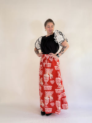 A person with tattooed arms stands against a light background, wearing a black top with sheer ruffled sleeves, black shoes, and the FARYLINA - FIFI WRAP SKIRT by FARYLINA featuring a white and beige cat pattern.