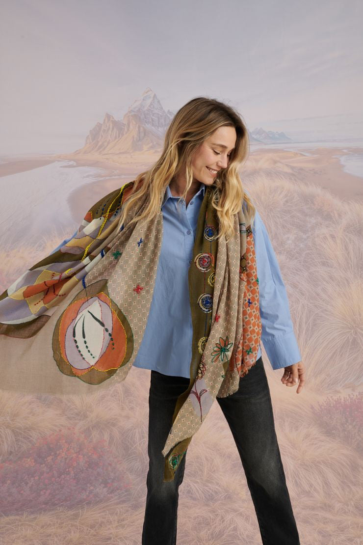 A woman with long blonde hair smiles at the camera in a scenic setting, wearing a white shirt, plaid pants, and the STORIATIPIC - ASTER WOOL SCARF by STORIATIPIC. Mountains, grass, and water are visible in the background.