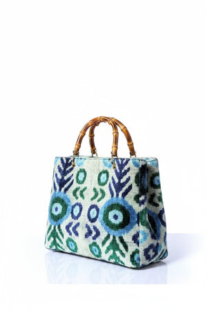 A person in light blue jeans and a white sleeveless top holds the BALDIZ AVERY Bag in Aqua Vine with bamboo handle. This stylish BALDIZ handbag’s blue, green, and white geometric design makes it fashionable and functional for any outing.