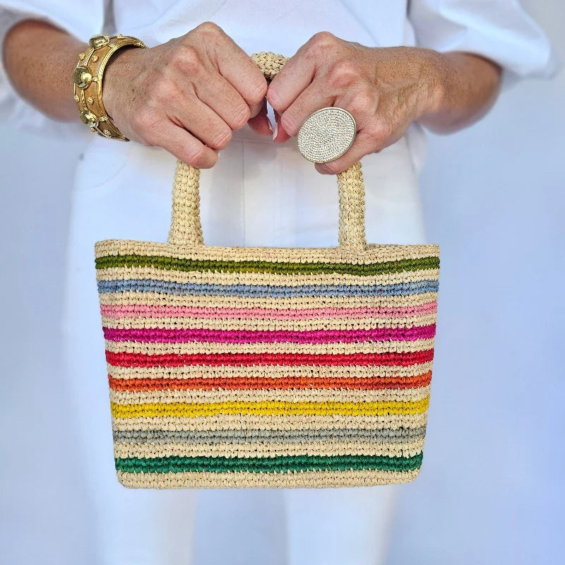 A person in white holds the BALDIZ MINI MAE RAFFIA BAG IN RAINBOW, an artisan-crafted tote with colorful stripes, styled with a chunky gold bracelet and a large, textured silver ring.
