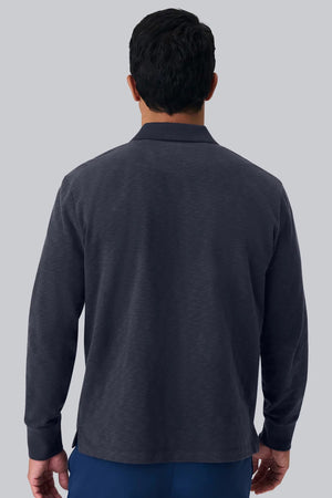 A man with short dark hair is seen from behind, wearing the MACK WELDON - HEAVYWEIGHT SLUB LONG SLEEVE POLO IN FROTH and blue pants, standing against a plain light gray background.