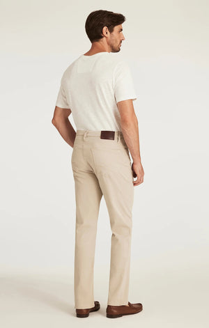 A man with light brown hair and a beard wears a tan blazer, brown sweater, white collared shirt, 34 HERITAGE - CHARISMA RELAXED STRAIGHT LEG PANT IN DAWN TWILL by 34 HERITAGE, and brown shoes against a plain white background.
