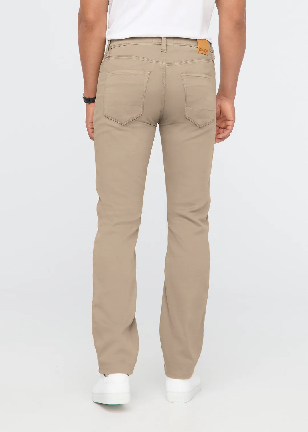 A man with short dark hair stands against a white background, wearing DUER No Sweat Straight Pants in Desert Khaki, a white t-shirt, and white sneakers. He faces the camera with a neutral expression.
