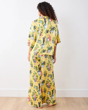 A person wears the PRINTFRESH - Macrame Mischief Satin Wildest Dream Pajama Set in Sunshine, featuring a vibrant cat and hanging plant pattern; their face is partially out of frame as they gently hold the shirt near the collar.