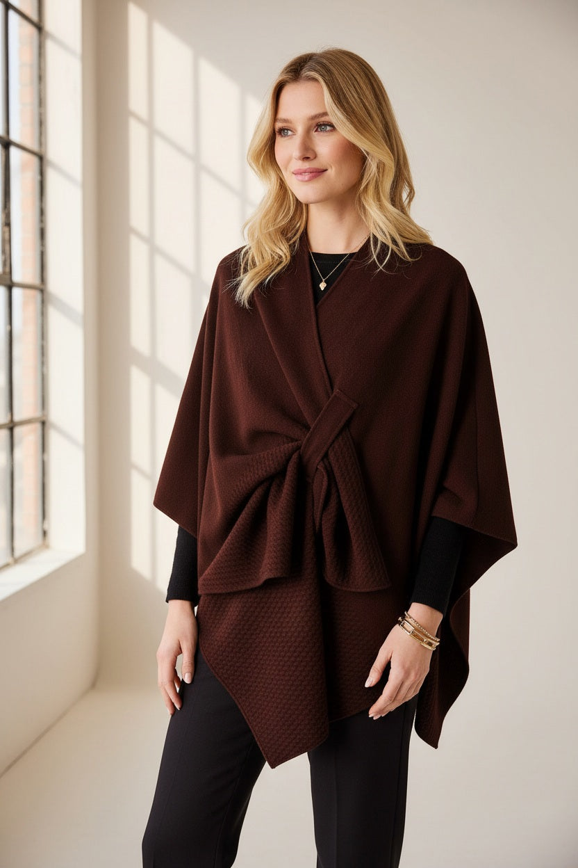 A woman stands against a plain background, wearing the MERSEA - Modern Travel Wrap in Chocolate with its convenient securing loop layered over her white top and loose brown pants. She completes the look with long blond hair and coordinating brown shoes.