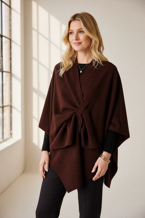 A woman stands against a plain background, wearing the MERSEA - Modern Travel Wrap in Chocolate with its convenient securing loop layered over her white top and loose brown pants. She completes the look with long blond hair and coordinating brown shoes.