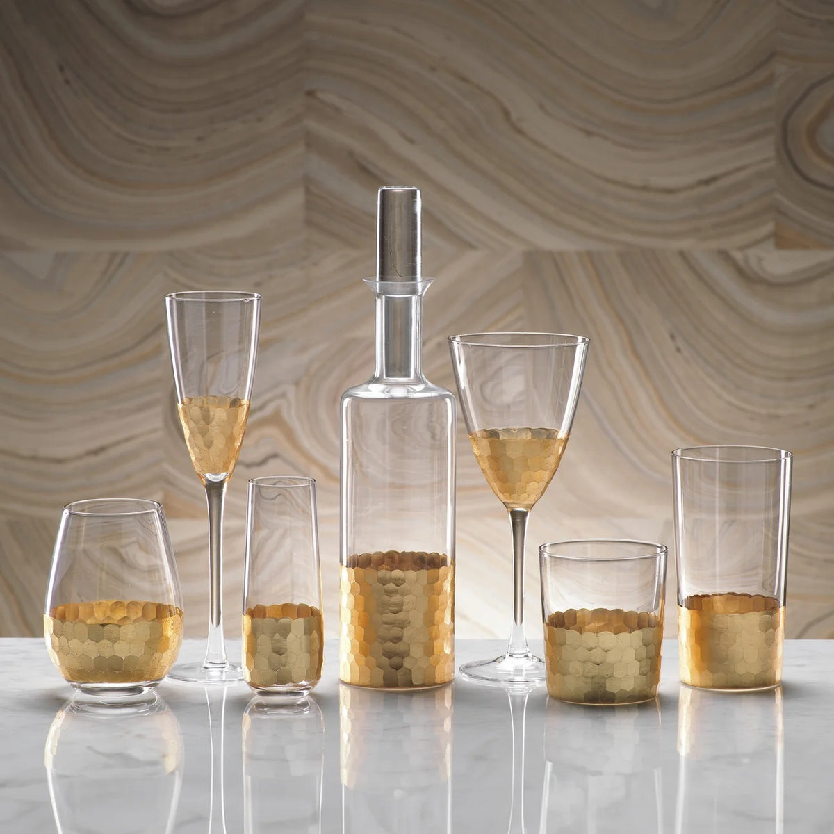 A collection of six glass vessels, including FEZ CUT STEMLESS CHAMPAGNE GLASS WITH GOLD LEAF, flutes, and a glass bottle decanter, each featuring a textured gold leaf honeycomb pattern at the base.