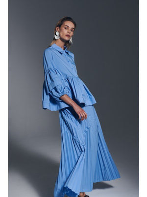 A woman models the MILVA MI Cotton Pleated Skirt in Azzurro Blue by MILVA, paired with a matching flowy top and voluminous sleeves, styled with bold white earrings against a plain gray background.