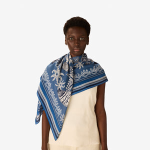 A person wearing a sleeveless white top styles the INOUI EDITIONS - NEPTUNE SCARF IN BLUE by INOUI around their neck, standing against a plain white background.