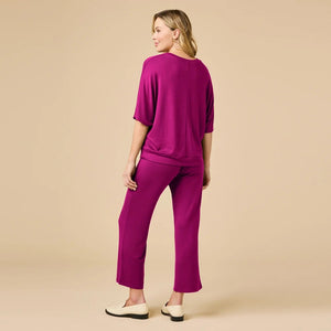 A woman wears a SOFTIES - DREAM RELAXED V-NECK WITH CAPRI LOUNGE SET in magenta, featuring a V-neck top with elbow-length sleeves and matching cropped drawstring pants, paired with cream-colored loafers.