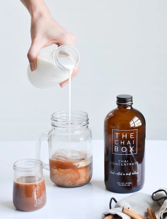 The Chai Box's brown glass bottle, prominently labeled with "THE CHAI BOX - CHAI CONCENTRATE," features text that reads "Hand crafted in small batches" and specifies a volume of 16 fl oz / 473 ml. Made with the finest spices, the label proudly displays "Atlanta, GA" at the bottom. The bottle is topped with a black screw cap.