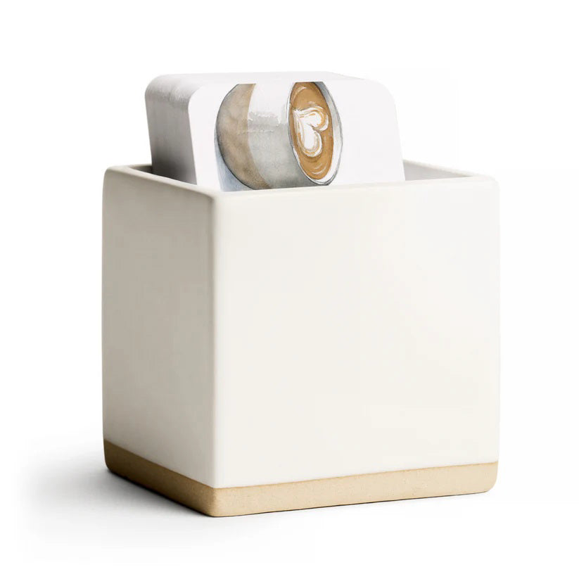 The E. FRANCES PAPER - LITTLE NOTES CERAMIC HOLDER - WHITE, designed as a modern minimalist ceramic cube gift box, contains decorative shredded paper and a gingerbread man-shaped white object, with the partially opened container revealing more shredded paper spilling out.